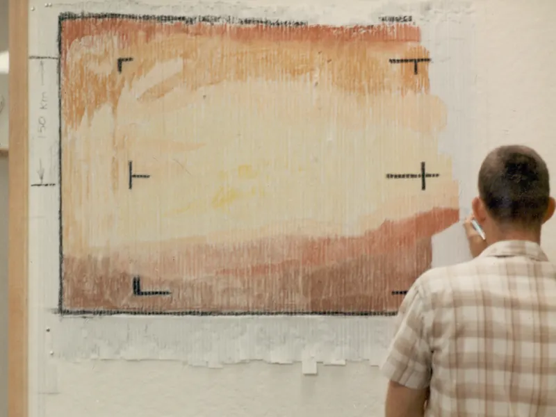 A large picture made of hand-coloured paper strips in orange and brown, on a wall, with a man looking at it