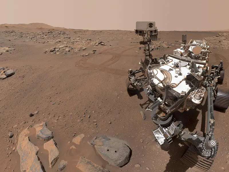 A six-wheeled rover on reddish ground taking a selfie, with a flat rock in front of it