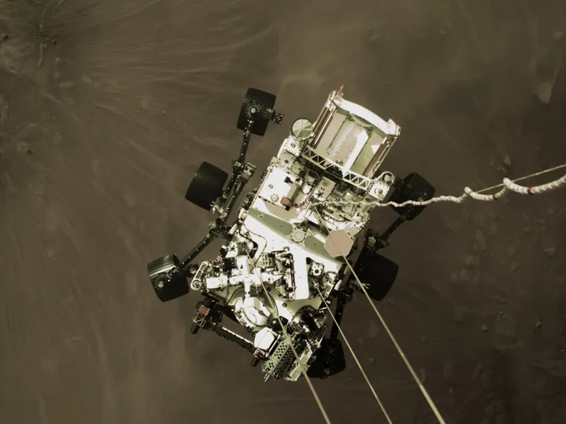 A six-wheeled rover hanging on cables below its descent stage, above dusty ground