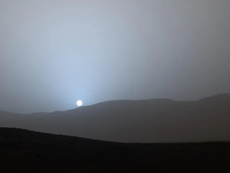 A dim blue glow around the setting Sun over the rim of a crater on Mars